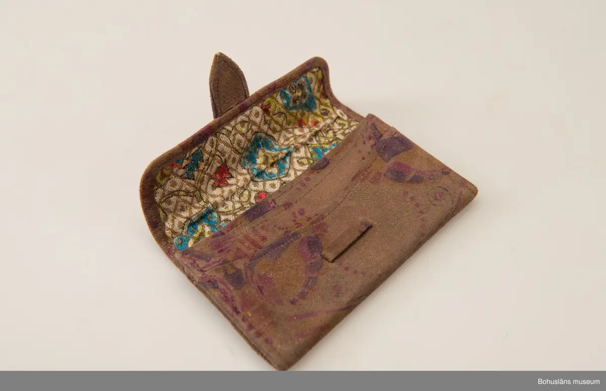 Brun börs av mockaskinn med tryckt blommönster i violett och rosa. Inuti blommönstrat foder på vit botten. Fyra fack.