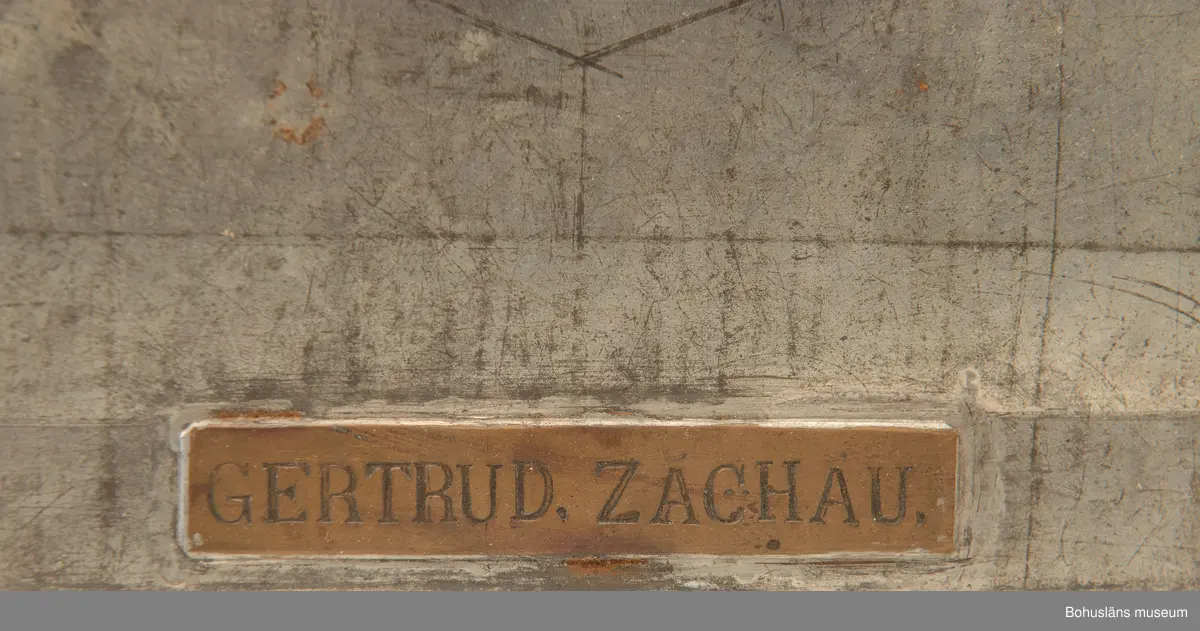 Rektangulär plåtlåda med lock. Handtag och nyckelhål. På lockets utsida en mässngsplåt med ingraverade texten GERTRUD ZACHAU.
Tillhörande nyckel.
Vid förvärvet innehöll skrinet fotogafier, se UMFA55947 och handlingar, se BMEA 488.