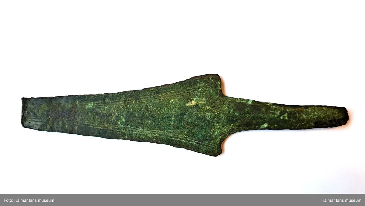 KLM 3387. Dolk, av brons. Med tvära skuldror och platt tånge. Klingan ornerad längs eggarna med tre enkla parallella fina linjer. Spetsen saknas.