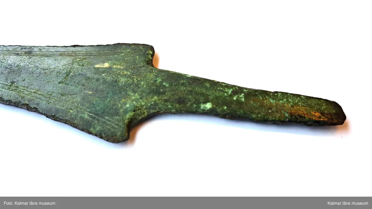 KLM 3387. Dolk, av brons. Med tvära skuldror och platt tånge. Klingan ornerad längs eggarna med tre enkla parallella fina linjer. Spetsen saknas.