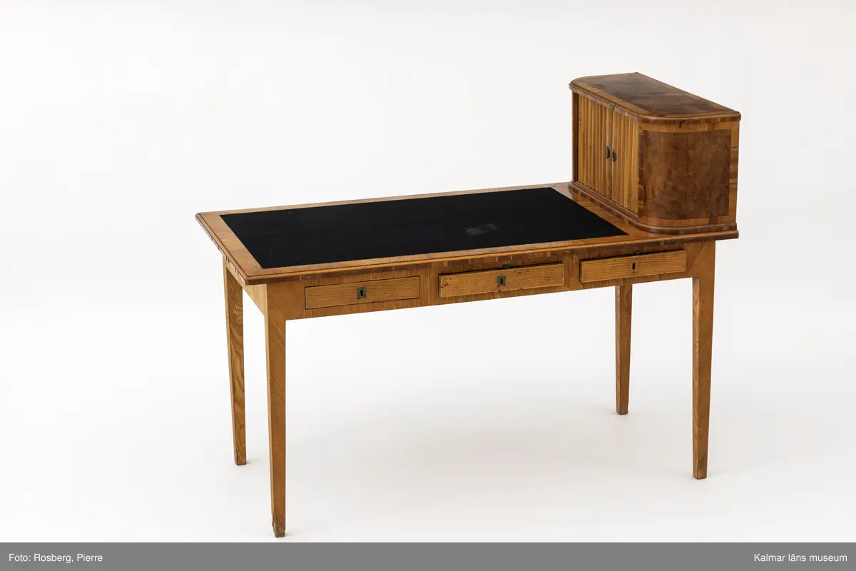 KLM 11136. Bord, skrivbord. Av trä med lövträdsfanér, eventuellt alm. Sengustavianskt med raka neråt avsmalnande fyrkantiga ben. Rektangulär skiva delvis klädd med svart filt samt längs ena långsidan tre lådor. Vid skivans kortände en hurts med jalusidörr, inuti två hyllplan samt tre små lådor.