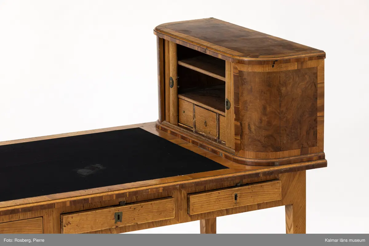 KLM 11136. Bord, skrivbord. Av trä med lövträdsfanér, eventuellt alm. Sengustavianskt med raka neråt avsmalnande fyrkantiga ben. Rektangulär skiva delvis klädd med svart filt samt längs ena långsidan tre lådor. Vid skivans kortände en hurts med jalusidörr, inuti två hyllplan samt tre små lådor.