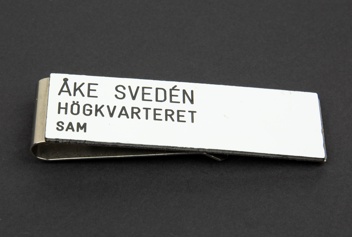 Namnskylt av typen klämma. På metallklämman är en vit etikett fastlimmad med texten "Åke Svedén Högkvarteret SAM"