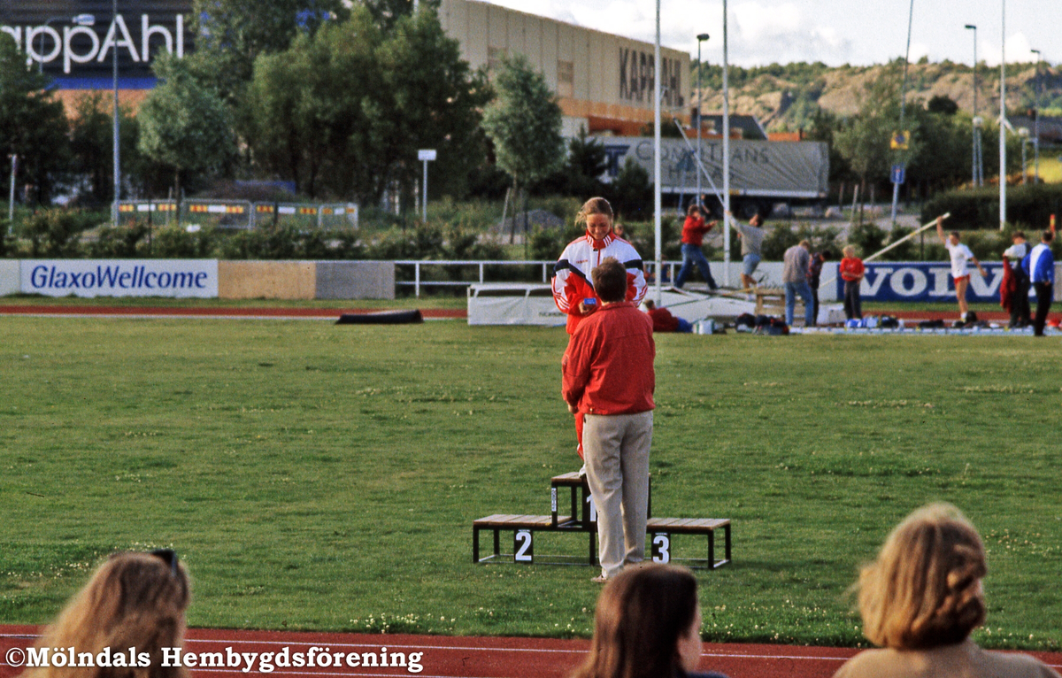Prisutdelning på Åby friidrottsstadion vid Idrottsvägen i Åby, Mölndal, den 18 juli 1996. En flicka står överst på prispallen och prisutdelare är Roger Boström från Mölndals-Posten. Anläggningen drivs av Mölndals AIK.
