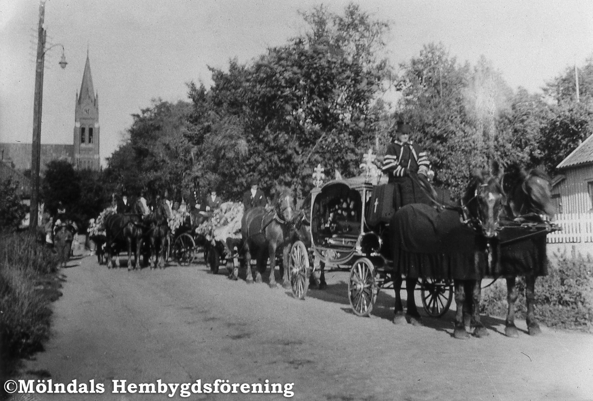 Albert Hedbergs begravningståg kör utefter Frölundagatan i Åby, Mölndal, med kyrkan i bakgrunden, september 1934. Albert var ägare till Katrinebergs gård. Han donerade även en summa pengar till Mölndals Hembygdsförening. AF 11:31.