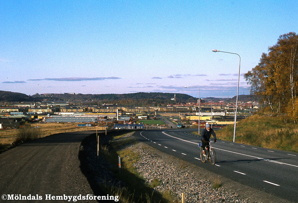 En cyklist cyklar uppför Pepparedsleden i Mölndal, år 1982. Åbro industriområde ses nedanför backen.
