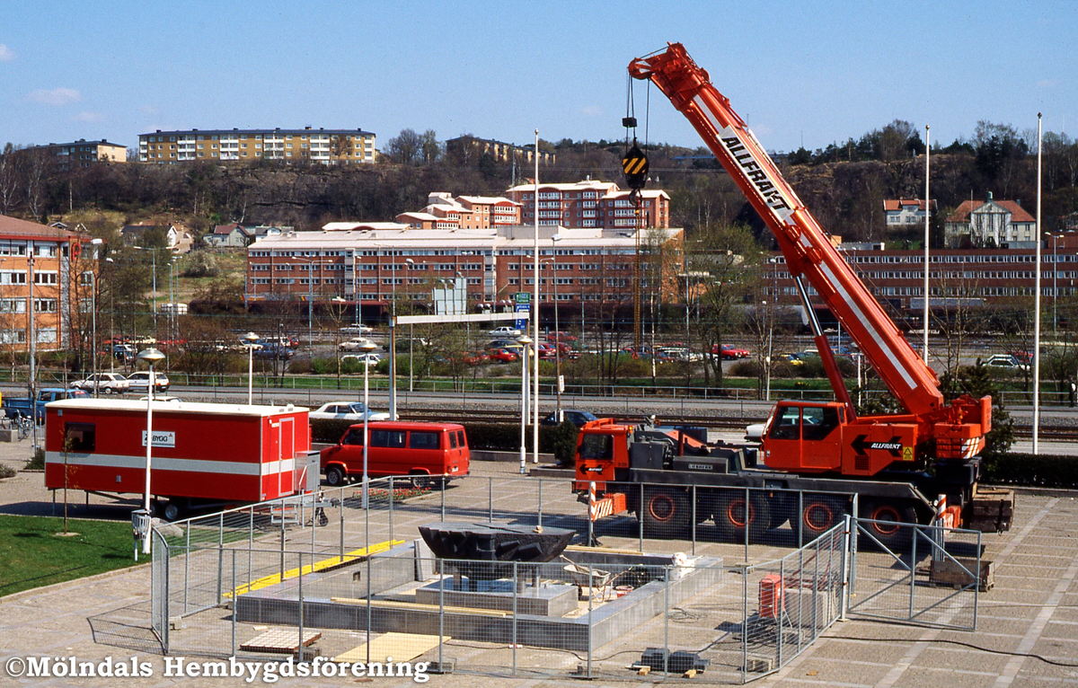 Konstverket Faesbiaergha av Roland Anderson installeras på Stadshusplan i Mölndal i maj 1991. Sockel och damm sätts på plats. I bakgrunden ses bebyggelse i stadsdelen Trädgården och upp på höjden i Enerbacken.