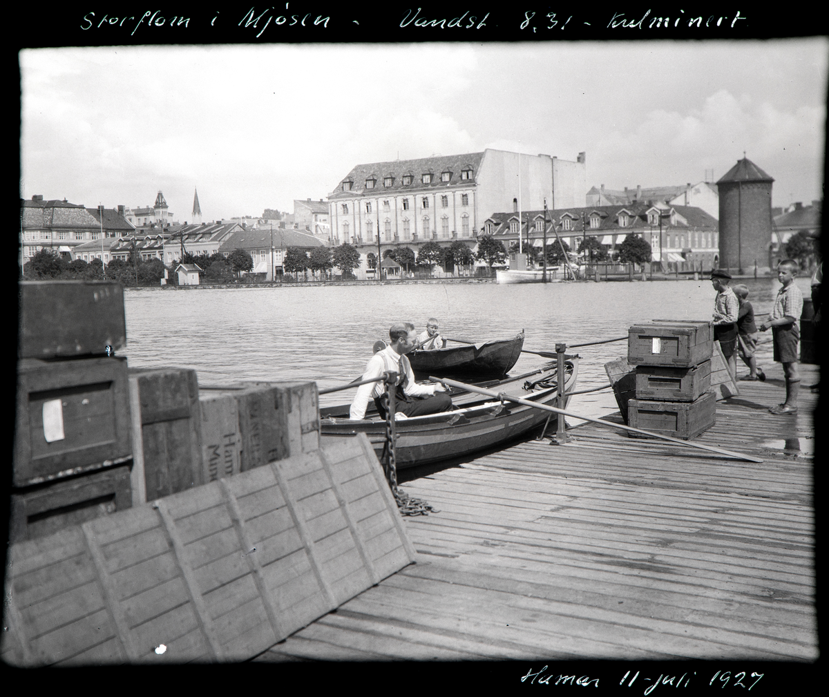 Hamar, Jernbanebrygga, Mjøsflommen 1927, robåter i Hamarbukta, vanntårnet, Festiviteten og Victoria hotell i bakgrunnen, jernbanespor, 11. juli 1927 vannstand 8,31 meter, i forgrunnen en robåt, bak en seilduksbåt med sønnen til fotografen: Hans Theodor Landstad (1916-),