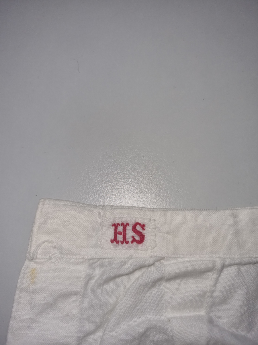 Bomull toskaft, hvit.
Åpning i sidene med trukket lerretsknapp.
Løpegang og trekksnor midt bak.
Nede på bena kapper av engelsk broderi.
Maskinsøm.
Merket: "H S"