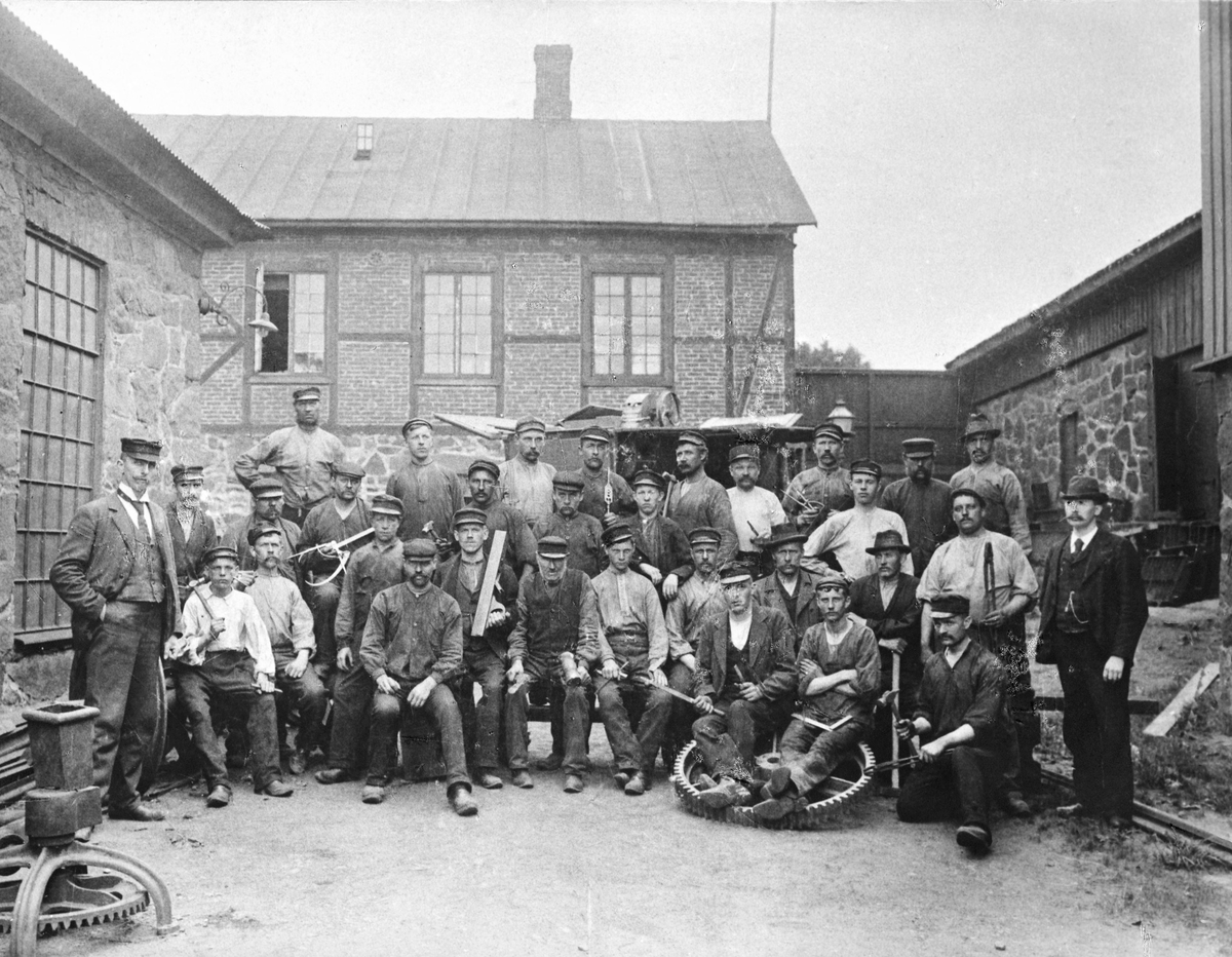 Arbetsstyrkan vid Varberg Mekaniska Verkstad omkring år 1900 samlad ute på gården. Med sig har männen olika verktyg som visar på deras profession; passare, tänger, klubbor, hammare, rubank med mera. Två män sitter dessutom på ett stort kugghjul. Här tillverkade man på beställning, främst redskap och maskiner som behövdes främst inom jordbruk och mejerinäringen men vissa produkter lagerhölls också. Förutom maskinavdelning fanns även snickeri- respektive plåtslageridel. Verkstaden startades på 1850-talet (då Ungewith & Co, spistillverkning) och drevs av olika personer fram till 1890-talet då Hugo Gerlach övertog företaget och brodern ingenjör Edmund Gerlach (död 1902) blev disponent. Verkstaden hade anlagts tämligen isolerat norr om den gamla trästaden, men vid det laget låg den mellan järnvägsstationen och den nya stenstaden, granne med de s k Tvillinghusen. Såsom ingenjörsdriven verksamhet fanns utöver gjuteri, maskinverkstad, smedja, magasin och koksbod även en ingenjörsbostad inom anläggningen. Enligt uppgift började arbetarna kl 6 och slutade 18.30. Maten fick de om vintern sitta och äta på ångpannan. (Se även bildnr VMA8141_1)

Sittande fr v: Skoog, Johan Svensson Bua, ?, Masen, Algot Svensson, Börje Ågren, Kastberg, Hillerström, Holmström, "Döven" Bengtsson, Hammarling, ?. Stående fr v: "Ingeniören" ( E. Gerlach?), Lövkvist, Maskinist Nilsson, Severin Svensson, Carlsson (farbror till boktryckare Carlsson), Nils på Lassabacka Andersson, ?, Julius Svensson, Gustaf Andersson, ?, Plåtslagare Carlsson, Eliasson Träslöv, Kalle Nia Karlsson, Söderlund, Gjutare Karlsson Träslöv, Bengtsson bror till Elisabeth Svensson, Kontorist Bengtsson (i hatt).