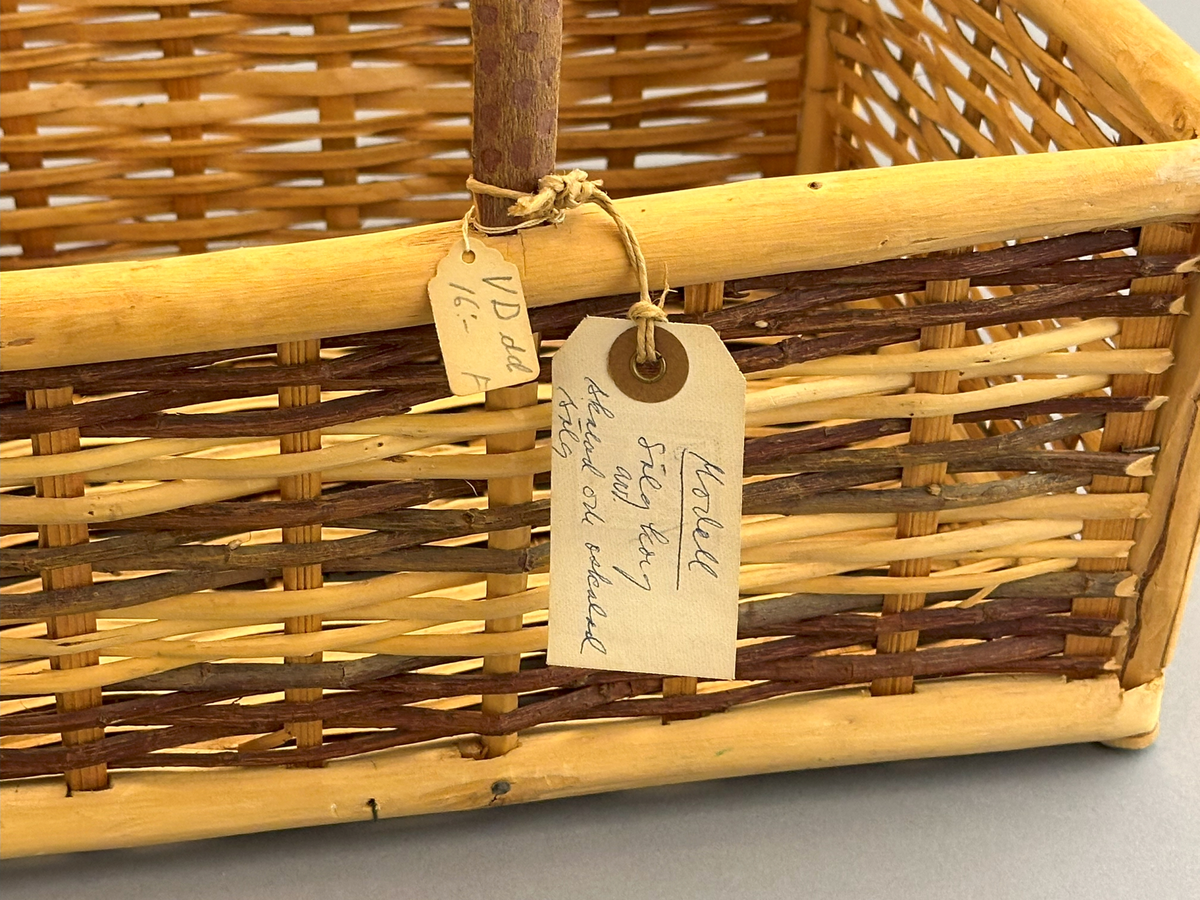 Fyrkantig korg med grepe, bestående av väggar både av skalad och oskalad sälg flätade i tuskaft på stående varp, insatta i ett ramverk av skalad sälg.