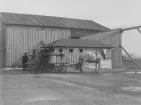 En häst med vagn står framför en lada i Österrike-Ungern, omkring 1914-1918. På vagnen står ett cirkelformat kar. I bakgrunden står en man. På ladan finns en skult med texten 