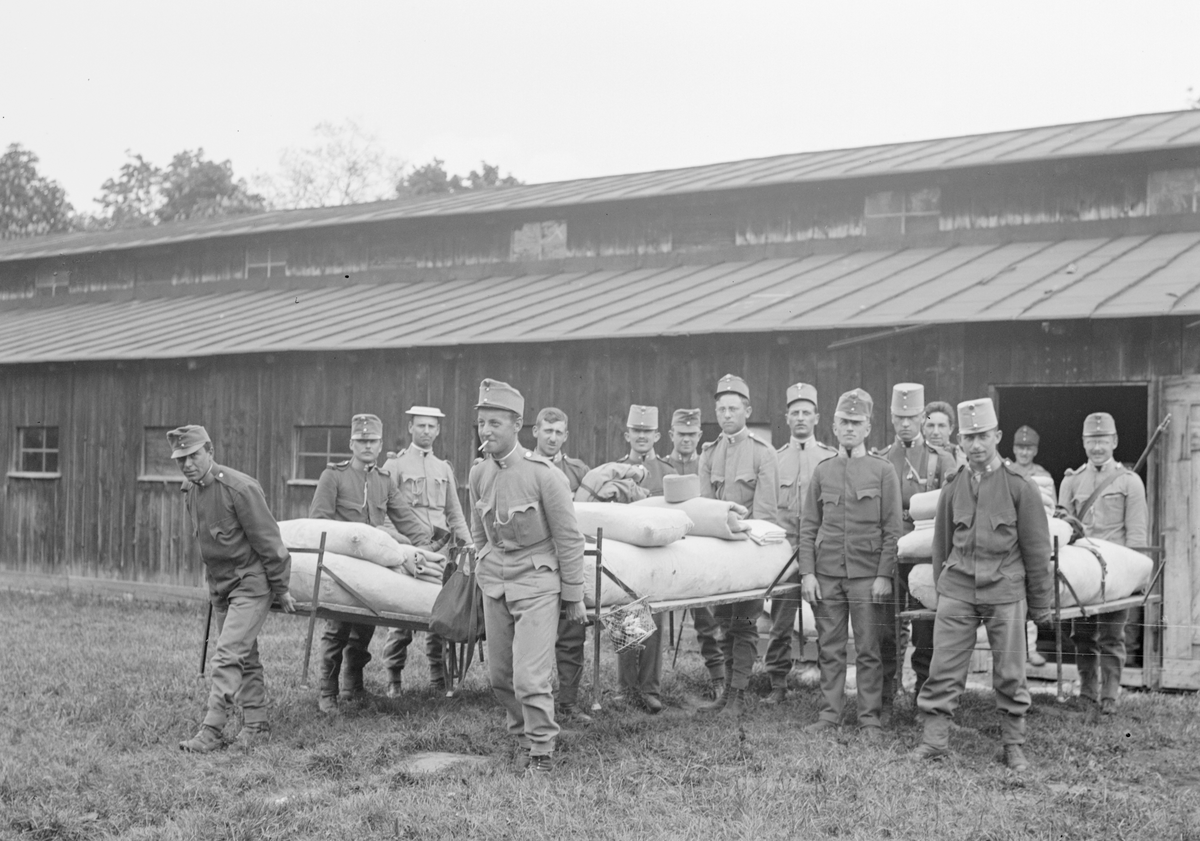 Militärer från Österrike-Ungern bär militärsängar utanför byggnad, 1914-1915. 15 militärer och tre sängar. Mannen i mitten, med cigarett i mun, är Edmund Sparmann.