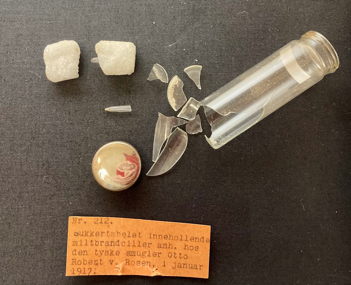 To sukkerbiter i glassylinder med metalllokk. Sylinderen er knust. Sukkerbitene inneholder svært små glassampuller.