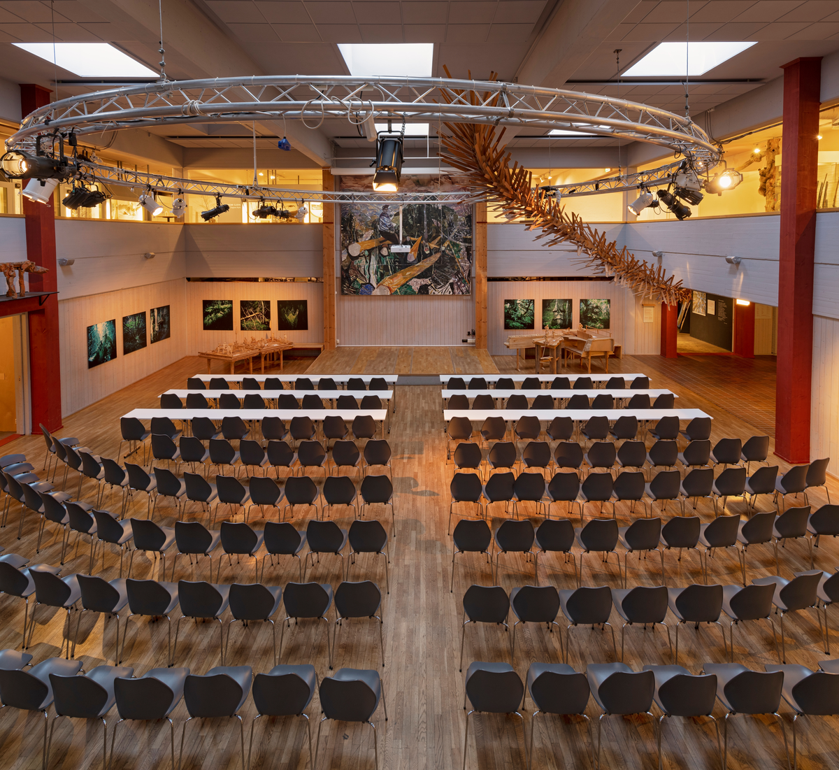 Fra sentralhallen i Norsk skogmusem i Elverum. Fotografiet ble tatt i januar 2022, i forkant av et møte der det var oppsatt stoler, og på de tre fremste rekkene også konferansebord, for et betydelig antall deltakere. I front, under Kai Fjells store maleri av en kvilende tømmerhogger, er det en 30 centimeter høy platting, der de opptredende skulle sitte eller stå. På begge sider av denne står det tablåer fra tømmerfløtinga i Osen i Åmot, utført av treskjæreren Ragnar Nysæther, og veggene er prydet med naturfotografier. Fra taket henger en rund lysrigg av aluminum. Fra denne henger det en treskulptur, utført av kunstneren Bo Jonzon, som peker ned mot inngangen til utstillingen «Tid for skog». Skogmuseets sentralhall fikk dette utseendet nettopp i forbindelse med bygginga av den nevnte basisutstillingen om skog og skogbruk, som ble bygd i periden 1999-2001. Rommet brukes leilighetsvis til konferanser og konserter, men står også ofte åpent, bare med noen få trebenker som innredning. Sentralhallen befinner seg i den sentrale delen av den første ildfaste museumsbygningen som ble reist på daværende Norsk Skogbruksmuseum i 1969-1970, etter tegninger av arkitekten Terje Thorstensen og med elverumsfirmaet Martin M. Bakken som hovedentreprenør. Bygningen ble innviet med kong Olav V som hedersgjest 22. juni 1971. Bygningen er seinere utvidet i to etapper (1979 og 1991-92).