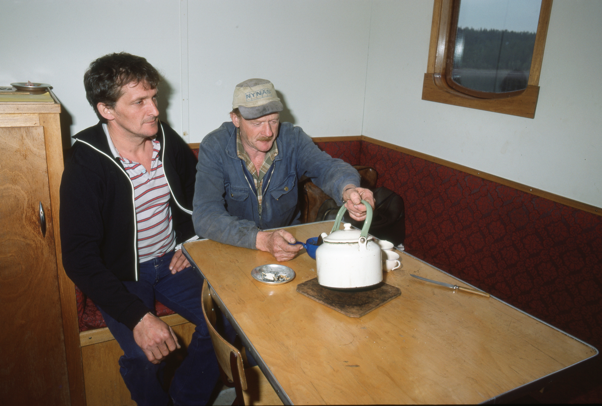 Fra salongen under dekk på slepebåten Trysilknut (Trysil-Knut). Fotografiet viser to av mannskapsmedlemmene, sittende på en vinklet benk ved et  bord med askebeger, kaffekopper og - kjele. Akkurat da dette fotografiet ble tatt var Nikolai Myrene, som var maskinist, i ferd med å skjenke kaffe fra en kvitemaljert kjele. Dette fotografiet ble antakelig tatt i 1984, den siste sesongen det foregikk tømmerfløting på Osensjøen. Denne innsjøen er om lag 28 kilometer lang og bortimot 3 kilometer bred. Slepebåten Trysilknut ble kontrahert ved Glommens mekaniske Verksted i Fredrikstad med sikte på at man skulle kunne mekanisere bukseringa av tømmeret over det 47 kvadratkilometer store vannspeilet. Komponentene ble transportert med jernbanen til Rena stasjon på Rørosbanen, og derfra ble de kjørt videre til Nordre Osen på hestesleder. Der ble komponentene klinket og skrudd sammen, slik at fartøyet kunne sjøsettes så snart isen var gått i 1914. Fra starten av hadde Trysilknut dampmaskin med en yteevne på 75 hestekrefter. Vinteren 1957-58 ble fikk båten dieselmotor med 135 hestekrefter. Samtidig ble både eksteriøret og interiøret en helt ombygd. Den nye innredningen, som vi ser noe av her, ble levert av Iver Hagens båtbyggeri på Hjelset ved Molde.