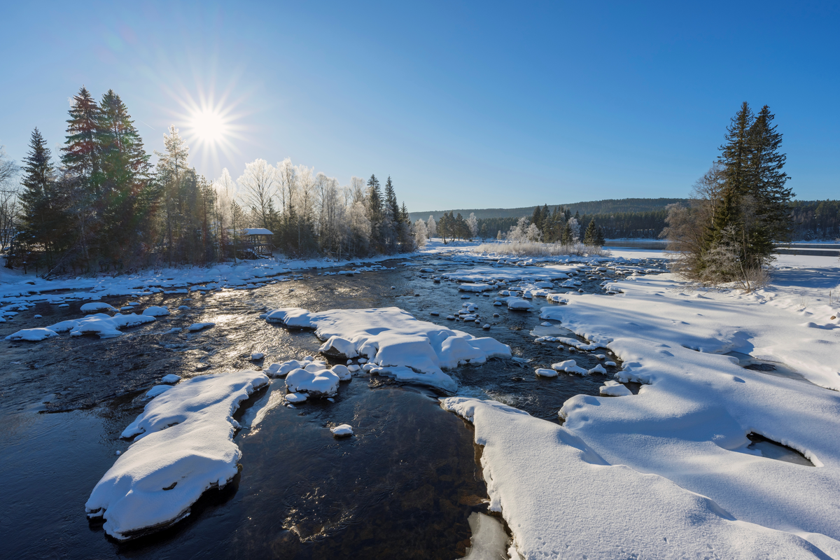 Fra nedre del av Klokkerfossen i Glomma i Elverum kommune i Hedmark, fotografert medstøms retning på en seinvinterdag i 2022, da vannføringa i elva var meget beskjeden. Bildet er tatt i motlys, som gir fine toner i vannet og i på snøen på de isdekte partiene i elveløpet. Klokkerfossen er betegnelsen på elvas østre løp forbi Prestøya, der det nordvestre løpet kalles Prestfossen. Klokkerfossen har antakelig fått dette navnet fordi gårdsbruket Fossum på østre elvebredd lenge var bosted for den av kommunens lærere som også skjøttet klokkerembetet i hovedkirka. I nyere tid har Norsk skogmuseum overtatt området. Ved den østre elvebredden (venstre halvdel av bildet) skimter vi Andrå-saga var Rendalen (SJF-B.0032), som ble flyttet til museet i 1963-64, og gjenoppført i elveskråningen. Den har måttet tåle en del påkjenninger i store flomår, og var nyrestaurert da dette fotografiet ble tatt. Sentralt i bildet ser vi ned mot Elverum camping.