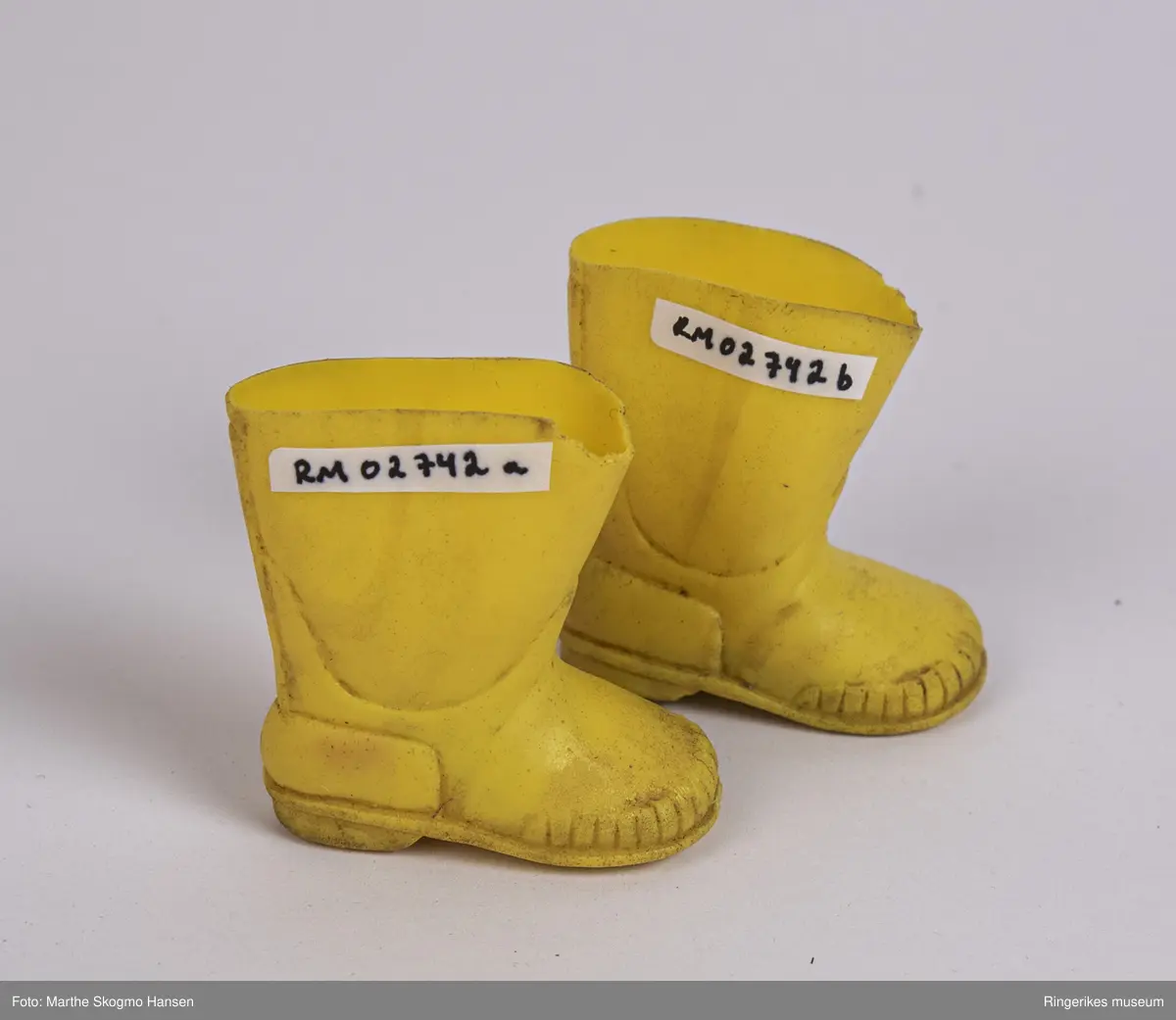 Gule gummistøvler til dokke. Støvlene er støpt i latex. På undersiden er det et merke hvor det står Latex, pluss tallet 6. Begge støvlene er litt slitt foran på skaftet, den ene (a) litt mer enn den andre. Hver støvel har en liten såle innvendig. Støvlene føles klissete når du tar på dem.