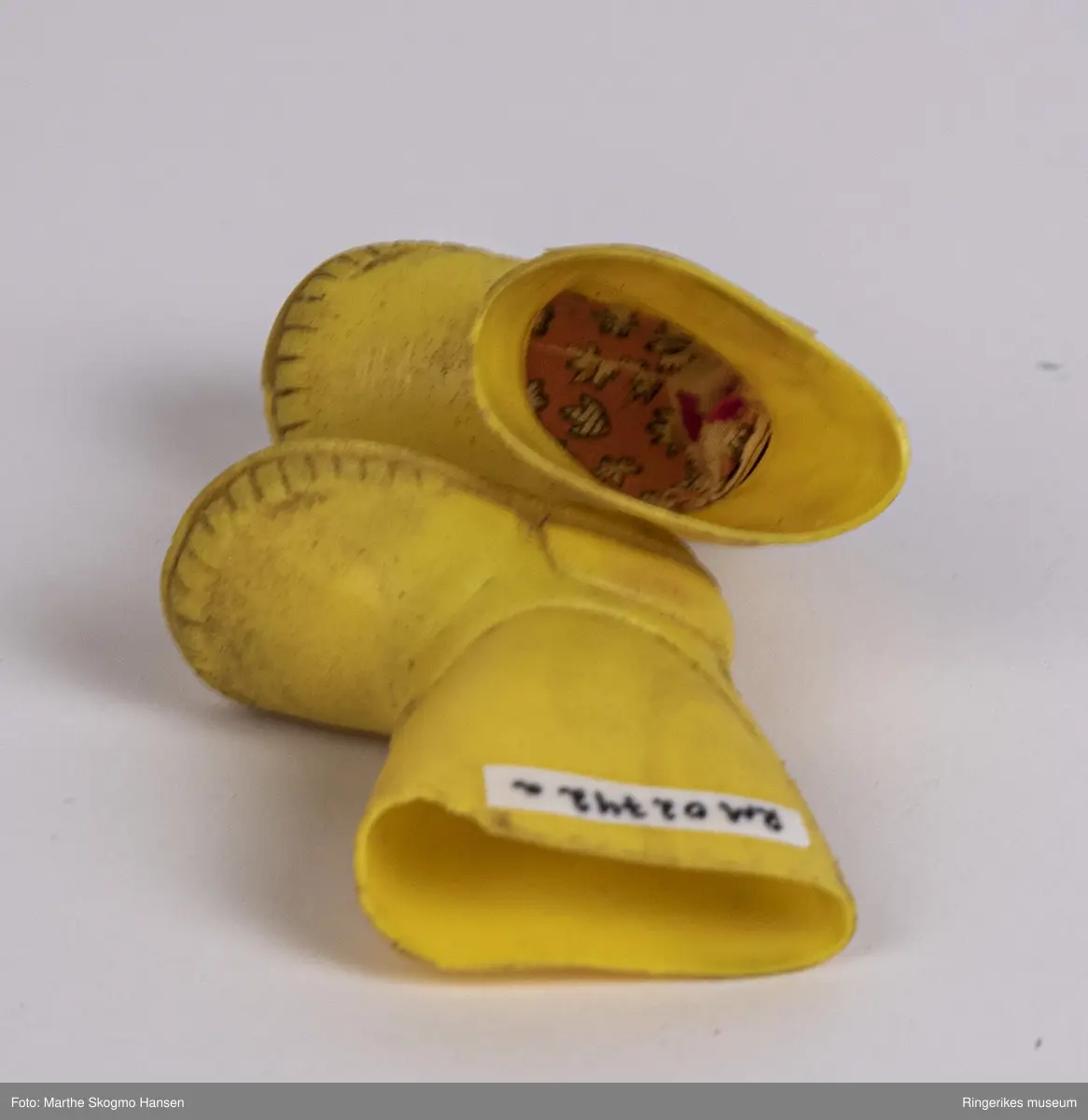 Gule gummistøvler til dokke. Støvlene er støpt i latex. På undersiden er det et merke hvor det står Latex, pluss tallet 6. Begge støvlene er litt slitt foran på skaftet, den ene (a) litt mer enn den andre. Hver støvel har en liten såle innvendig. Støvlene føles klissete når du tar på dem.