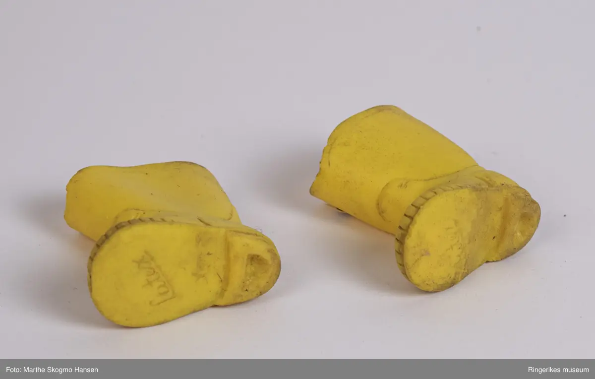 Gule gummistøvler til dokke. Støvlene er støpt i latex. På undersiden er det et merke hvor det står Latex, pluss tallet 6. Begge støvlene er litt slitt foran på skaftet, den ene (a) litt mer enn den andre. Hver støvel har en liten såle innvendig. Støvlene føles klissete når du tar på dem.