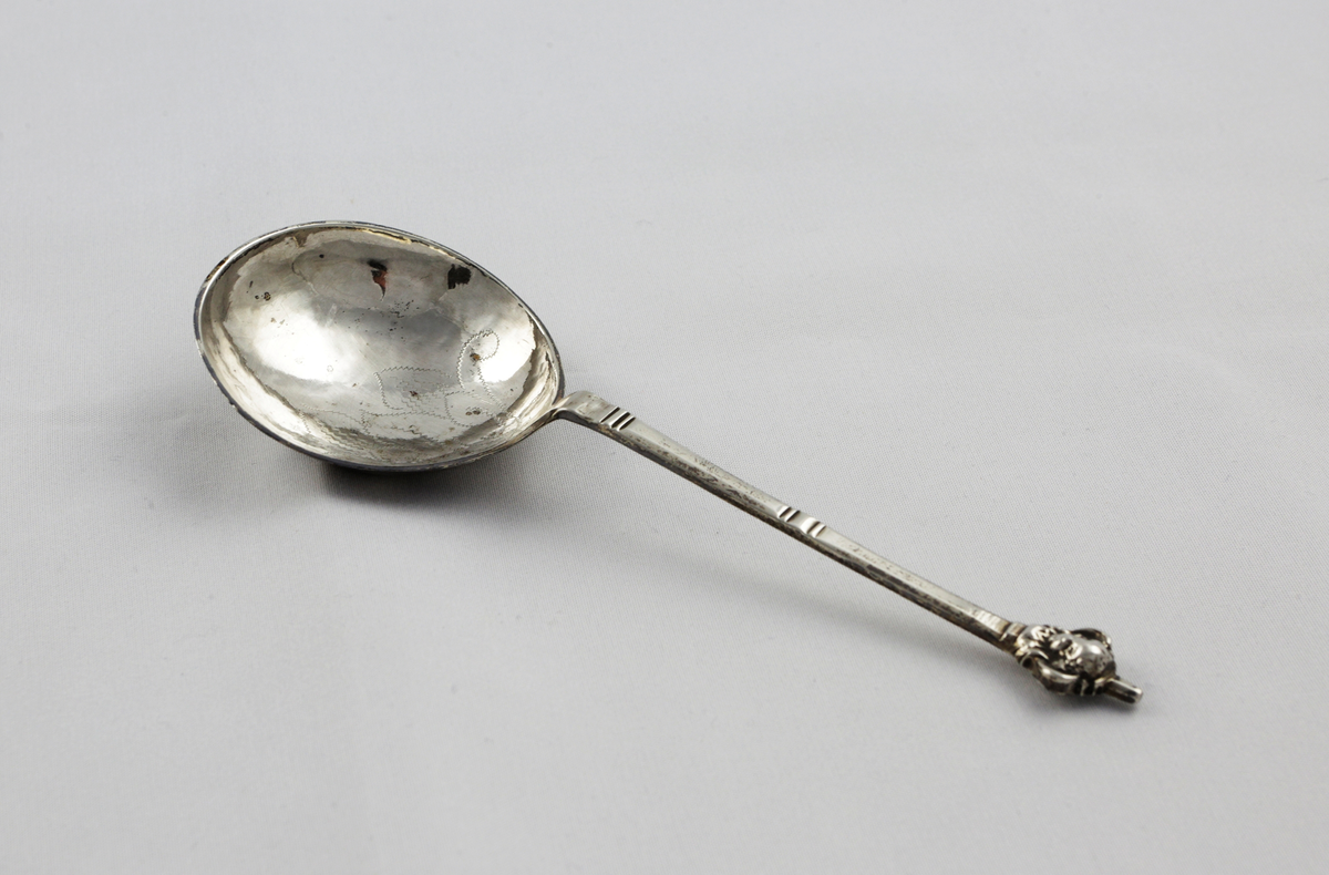 Brännvinssked i ett stycke med runt skedblad. Graverat mönster med blomstermotiv. Fyrsidigt skaft med tvärgående räfflor och kerubknopp.