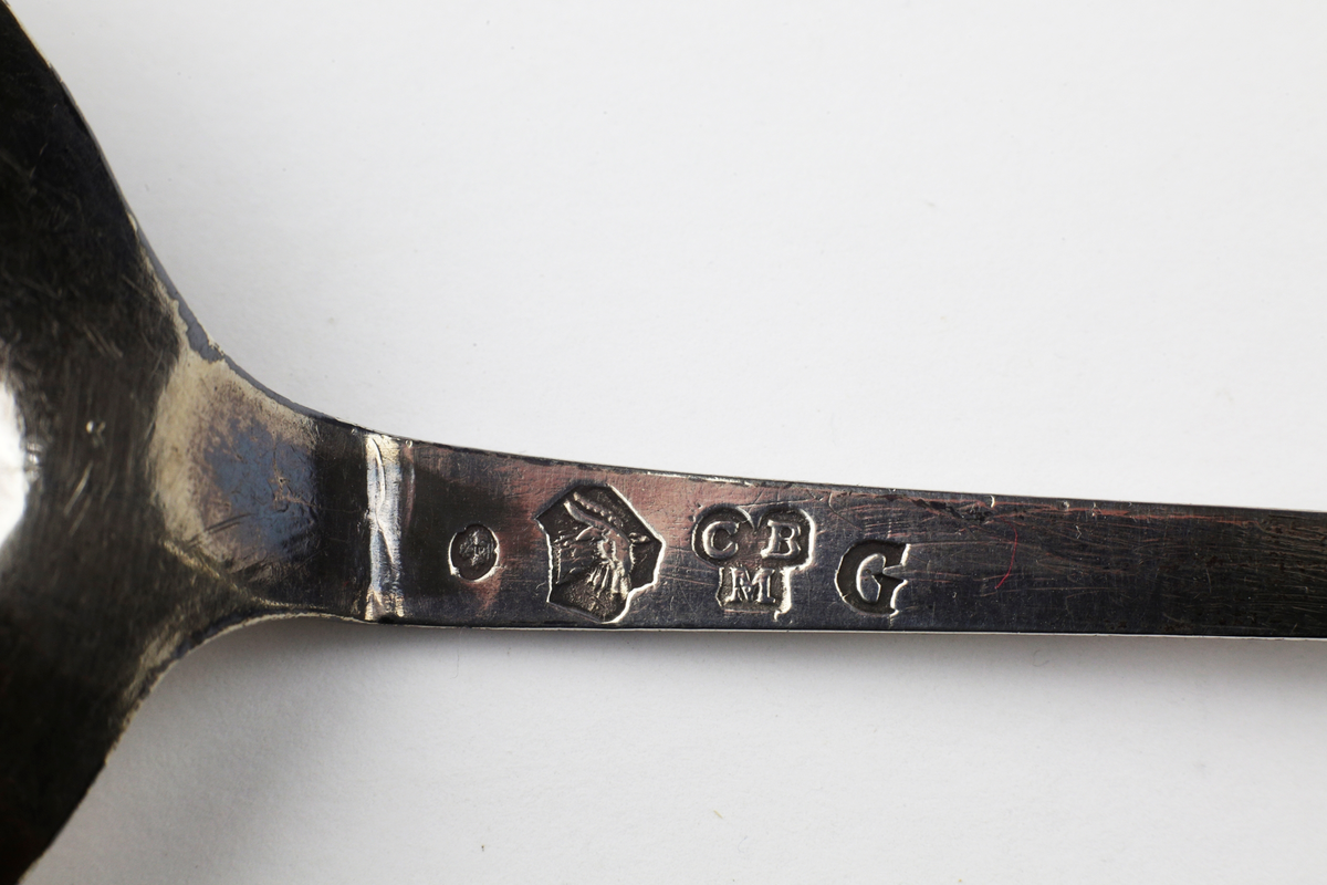 Brännvinssked i ett stycke med runt skedblad. Graverat mönster med blomstermotiv. Fyrsidigt skaft med tvärgående räfflor och kerubknopp.