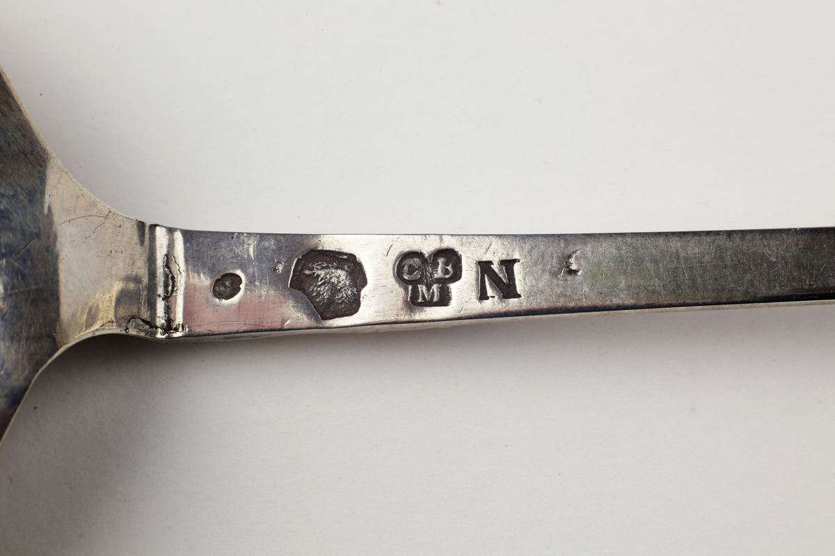 Brännvinssked med svagt päronformigt skedblad. Förgyllt graverat mönster med blomstermotiv. Skaftet avslutat med en förgylld kronknopp med fem små ringar.