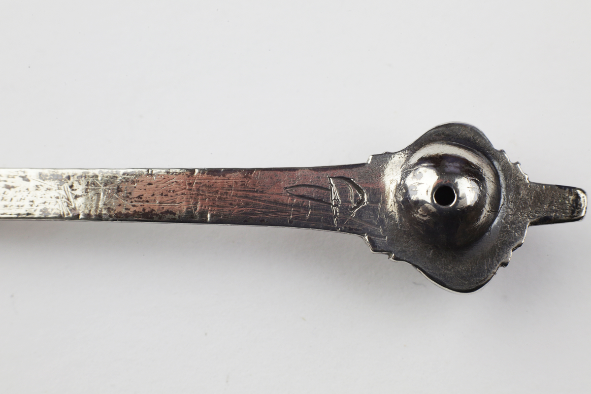 Brännvinssked i ett stycke med runt skedblad. Graverat mönster med blomstermotiv. Fyrsidigt skaft med tvärgående räfflor och kerubknopp.