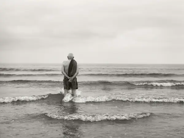En mann står ute i vannet, han har på shorts og har et håndkle over skuldra. Utsikten er kun hav.
