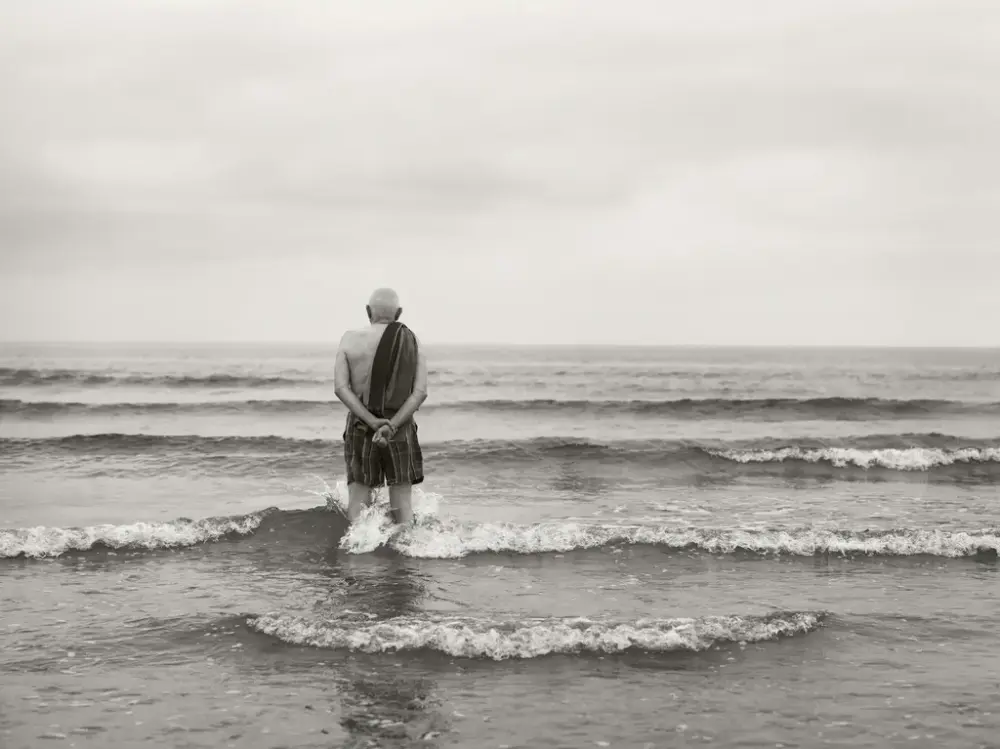 En mann står ute i vannet, han har på shorts og har et håndkle over skuldra. Utsikten er kun hav.