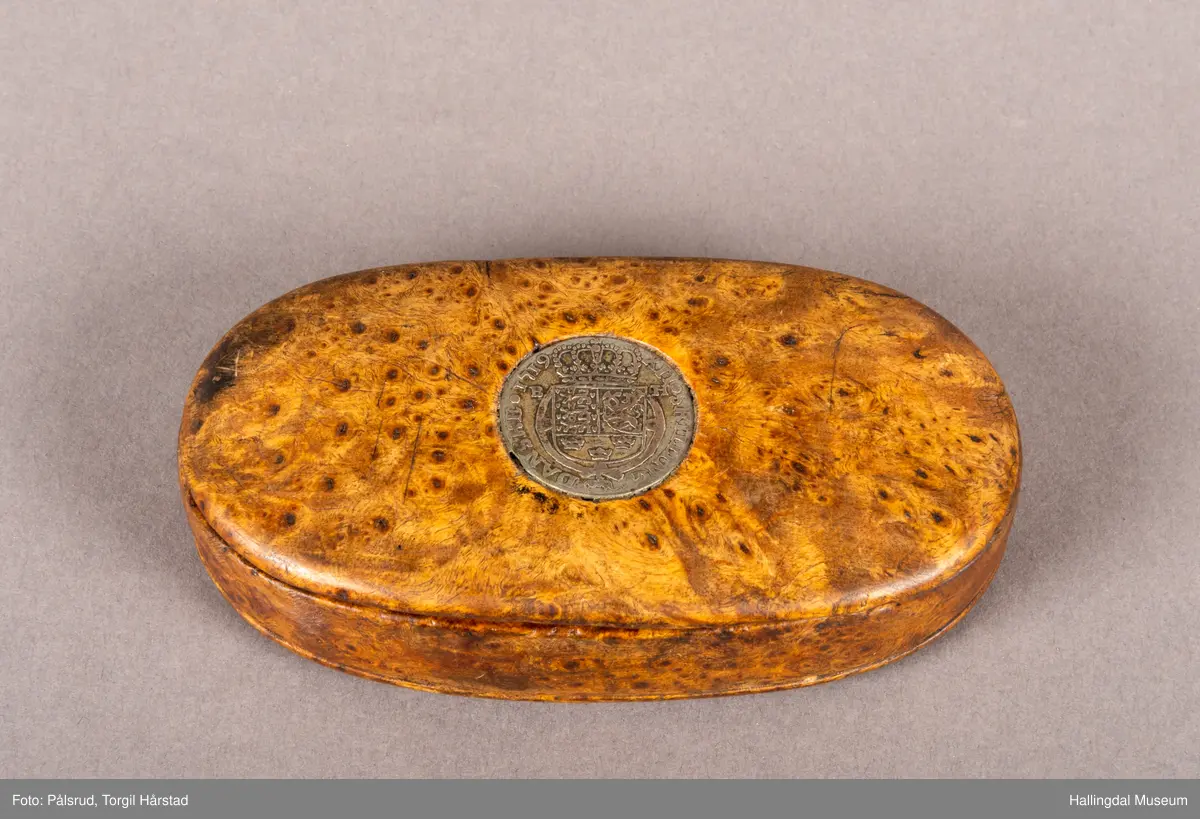 En brun, oval snusdåse i valbjørk. Lokket er festet med trehengsler på den ene langsiden. Innfelt i lokket sitter en 12-skilling fra 1719. Under bunnen er det innskåret "K O S ANO 1642".