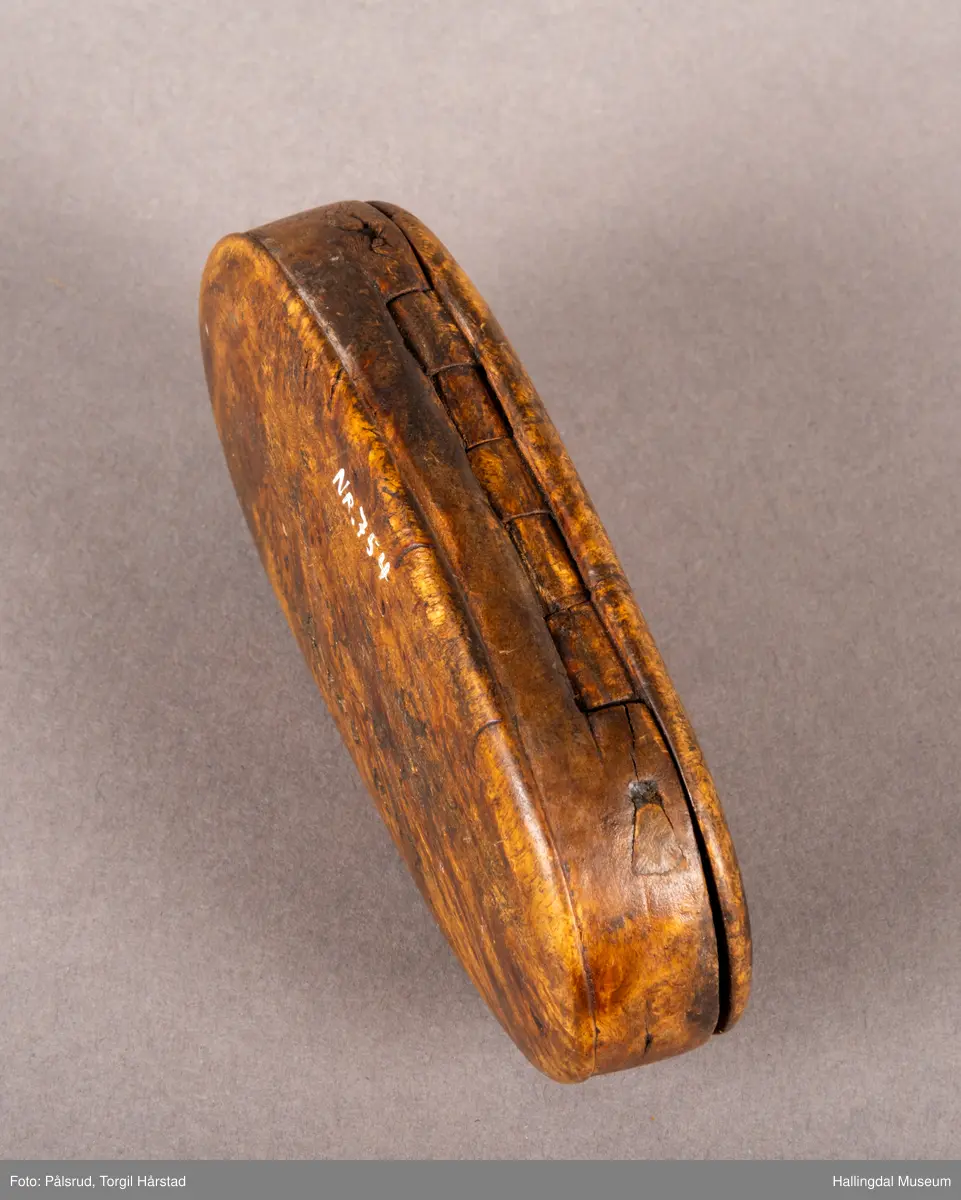 En brun, oval snusdåse i valbjørk. Lokket er festet med trehengsler på den ene langsiden. Innfelt i lokket sitter en 12-skilling fra 1719. Under bunnen er det innskåret "K O S ANO 1642".