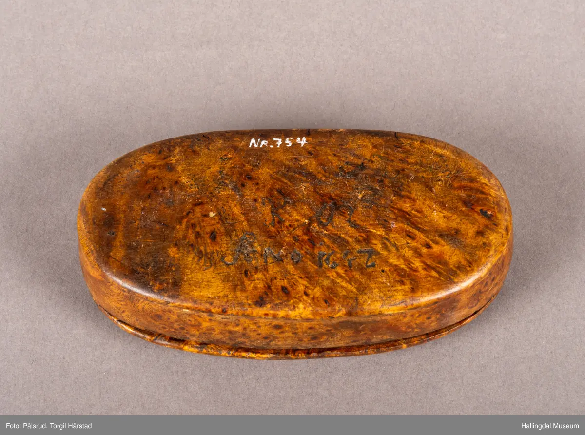 En brun, oval snusdåse i valbjørk. Lokket er festet med trehengsler på den ene langsiden. Innfelt i lokket sitter en 12-skilling fra 1719. Under bunnen er det innskåret "K O S ANO 1642".