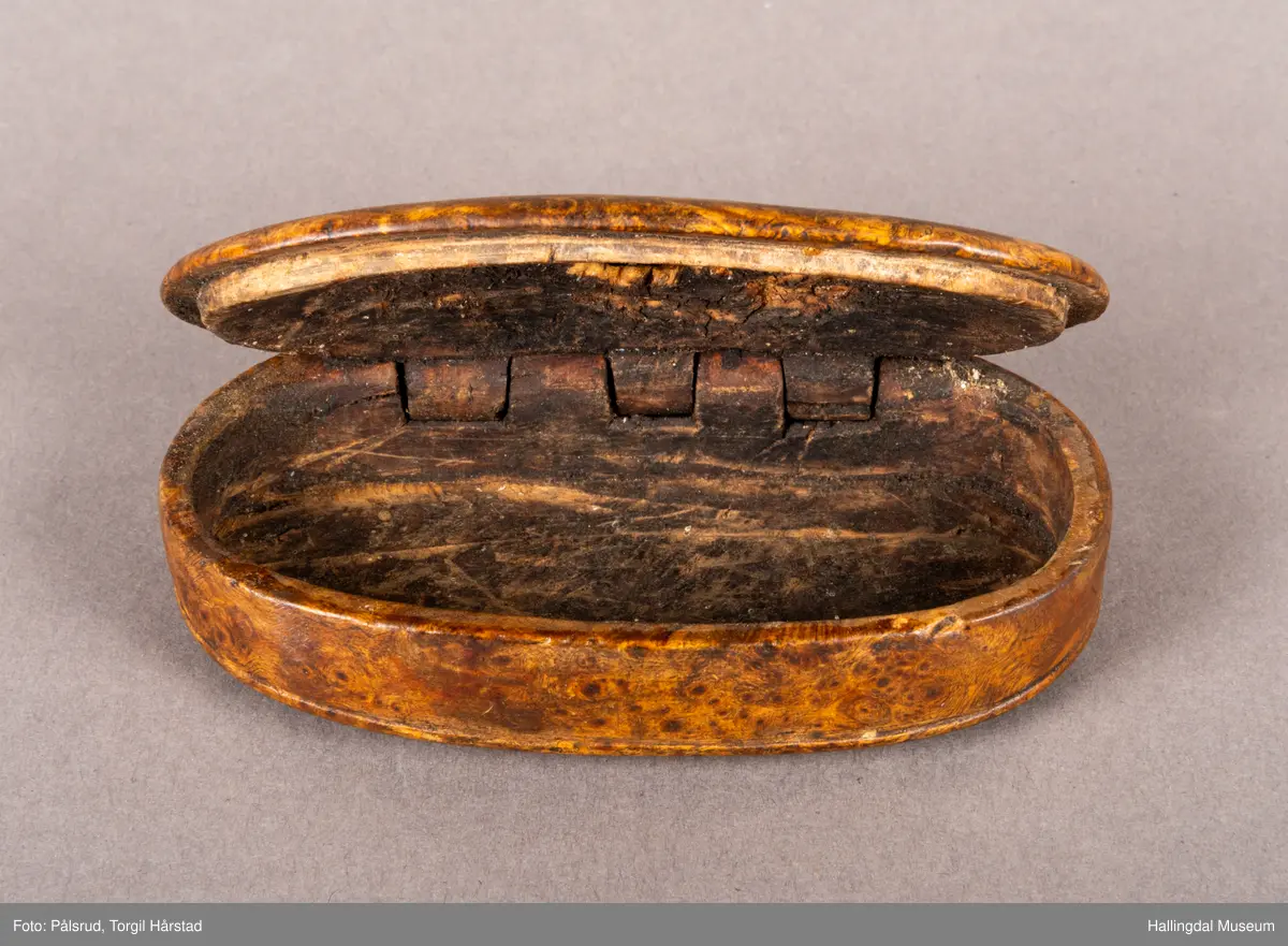 En brun, oval snusdåse i valbjørk. Lokket er festet med trehengsler på den ene langsiden. Innfelt i lokket sitter en 12-skilling fra 1719. Under bunnen er det innskåret "K O S ANO 1642".