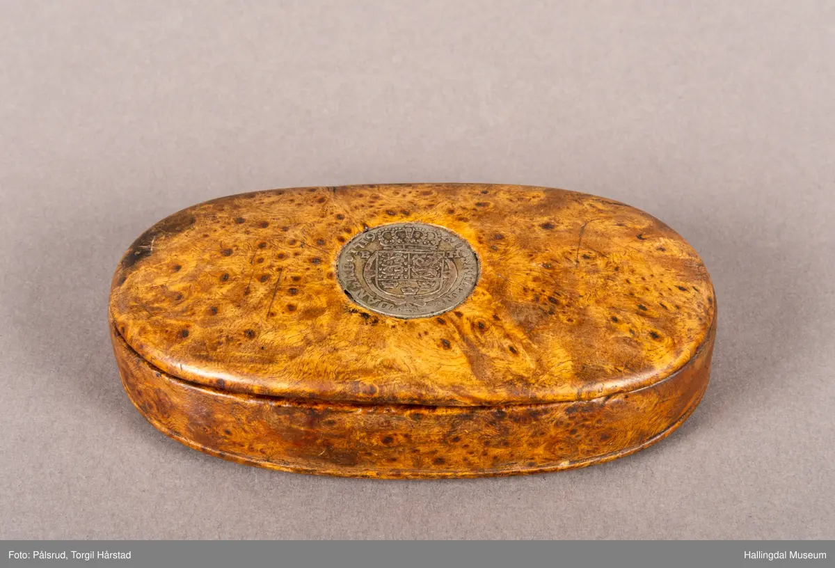 En brun, oval snusdåse i valbjørk. Lokket er festet med trehengsler på den ene langsiden. Innfelt i lokket sitter en 12-skilling fra 1719. Under bunnen er det innskåret "K O S ANO 1642".