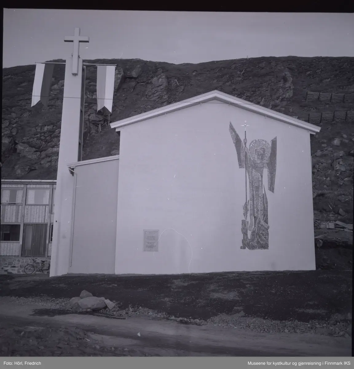 Ved klokketårnet til den katolske kirka St. Mikael i Hammerfest er det montert to stenger med vimpler. Det antas at dette ble gjort i anledning vigslingen den 3. august 1958. 
Kirkens vegg mot gata er dekorert med en stor mosaikk av St. Mikael, skapt av den tyske billedkunstneren Karl Manninger. Ved kirkens sidefløy til venstre er en sykkel lent mot veggen. Bak kirkebygget ser man Salenfjellet.