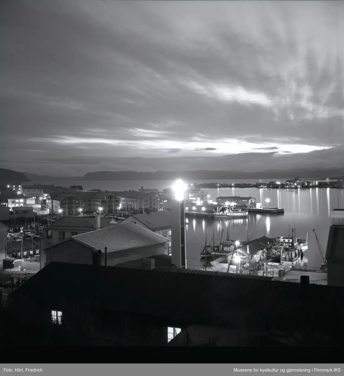 Det er skumringstime i Hammerfest og fotografen tar et bilde fra fjellsiden av Salen ut mot havna. I forgrunnen ser man ei brakke, der det er lys i to av vinduene. Bak brakka ser man det lyssatte korset ved klokketårnet til den nybygde katolske kirka St. Mikael. Gatelysene er på. Ved kaia ligger det båter og på andre siden av bukta ser man bydelen Fuglenes. Lengst bak i bildet ser man silhuetten av Sørøya.