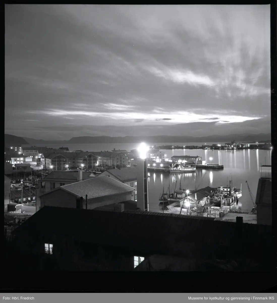 Det er skumringstime i Hammerfest og fotografen tar et bilde fra fjellsiden av Salen ut mot havna. I forgrunnen ser man ei brakke, der det er lys i to av vinduene. Bak brakka ser man det lyssatte korset ved klokketårnet til den nybygde katolske kirka St. Mikael. Gatelysene er på. Ved kaia ligger det båter og på andre siden av bukta ser man bydelen Fuglenes. Lengst bak i bildet ser man silhuetten av Sørøya.