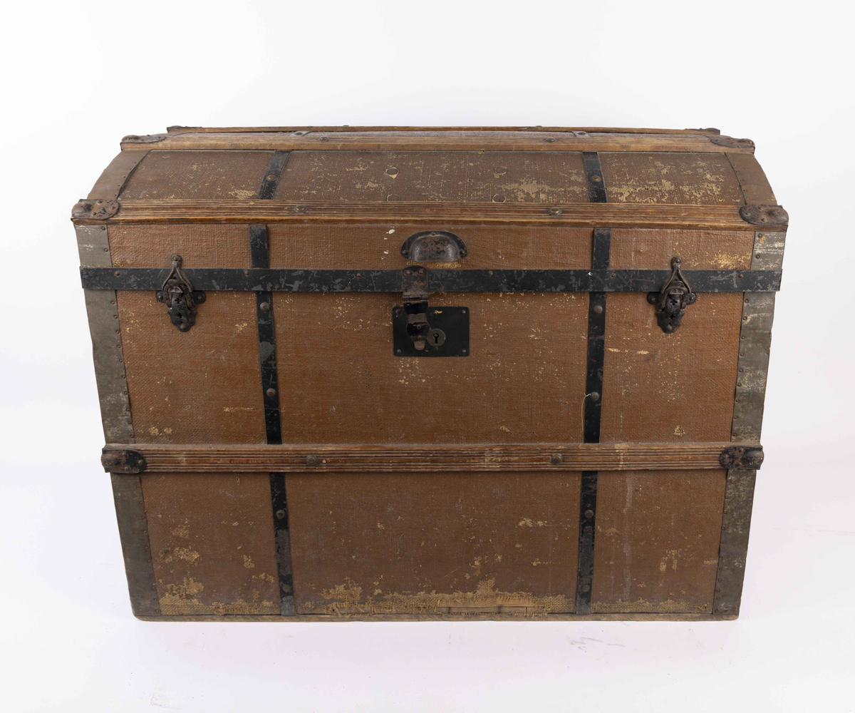 Stor amerikakoffert med buet lokk. Den er kledd med brunmalt lerret festet med trelister og svarte metallbeslag. Hver kortside har en metallhank, og fremsiden har tre metallåser. Innsiden er kledd med stripete papirtapet.