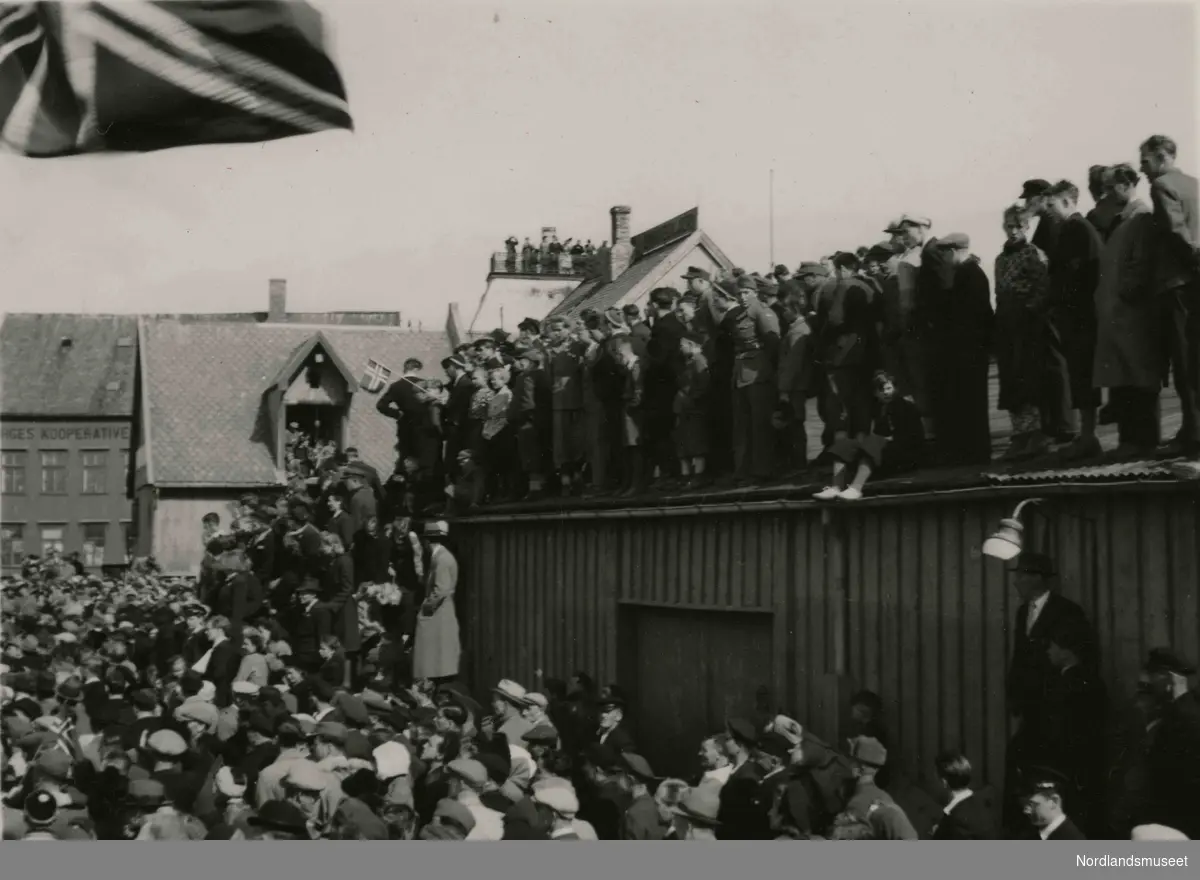 En folkemengde. Mange står oppå et blikktak. Bilde tatt i Bodø under feiring av freden etter okkupasjonen 1945.