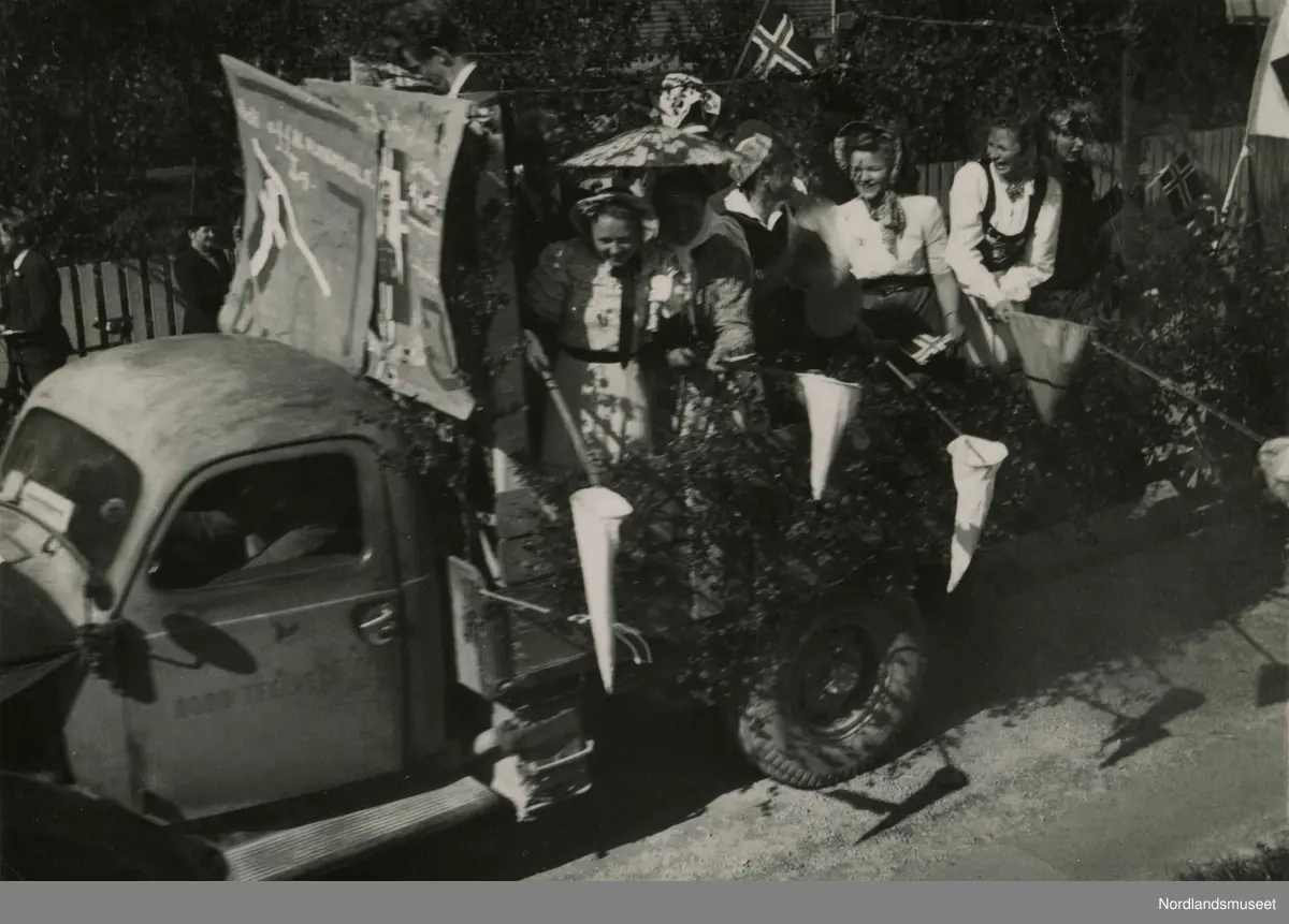 En lastebil pyntet med bjørkekvister. I lasterommet står det unge kvinner i pene klær. De holder noe som ligner på håver. Bilde tatt i Bodø under feiring av freden etter okkupasjonen 1945.