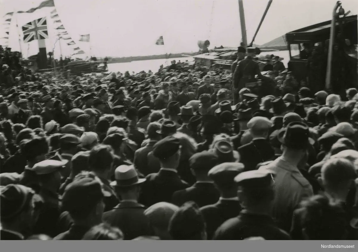 En folkemengde. Et skip i bakgrunnen med britisk flagg. Bilde tatt i Bodø under feiring av freden etter okkupasjonen 1945.