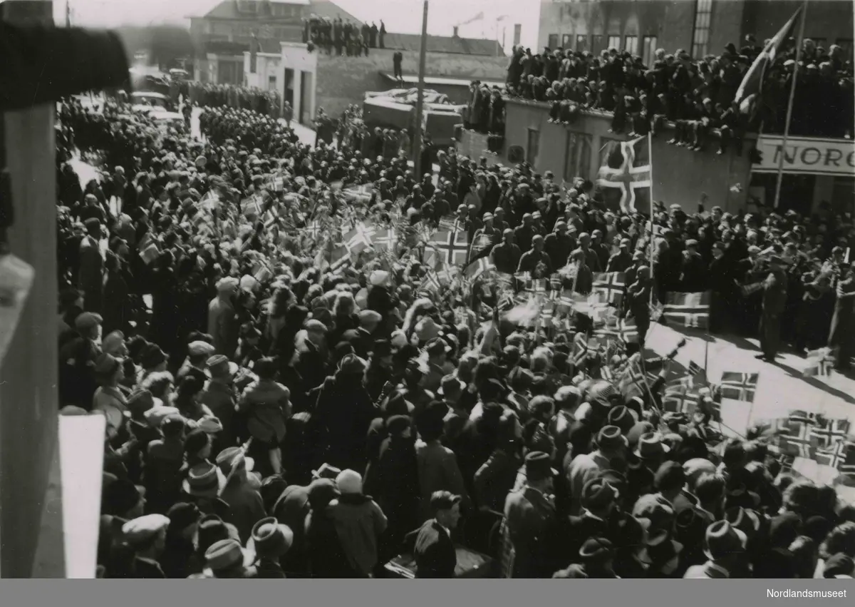 En folkemengde i en bygate. Noen sitter på tak. Bilde tatt i Bodø under feiring av freden etter okkupasjonen 1945.