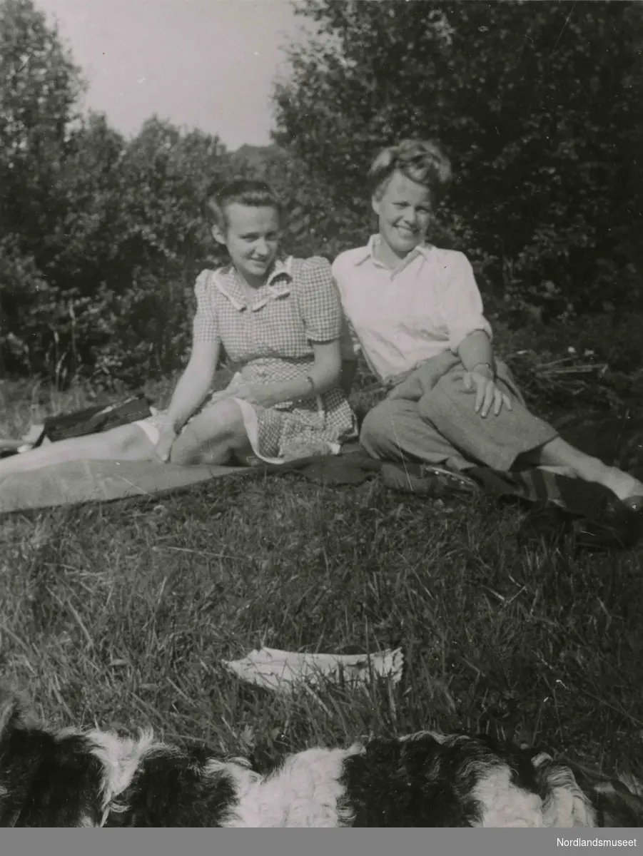 To unge kvinner sitter på gresset. En hund ligger i forgrunnen. Solveig Simonsen (gift Hunstad) til venstre.