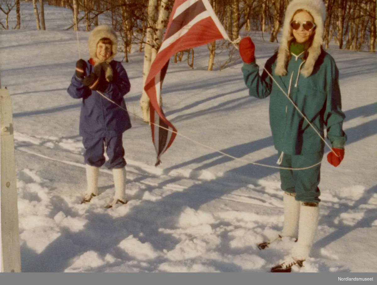 To barn holder en flaggvimpel. Det er vinter. De har på seg vinterklær og beksømsko. Øyunn og Ingvild.