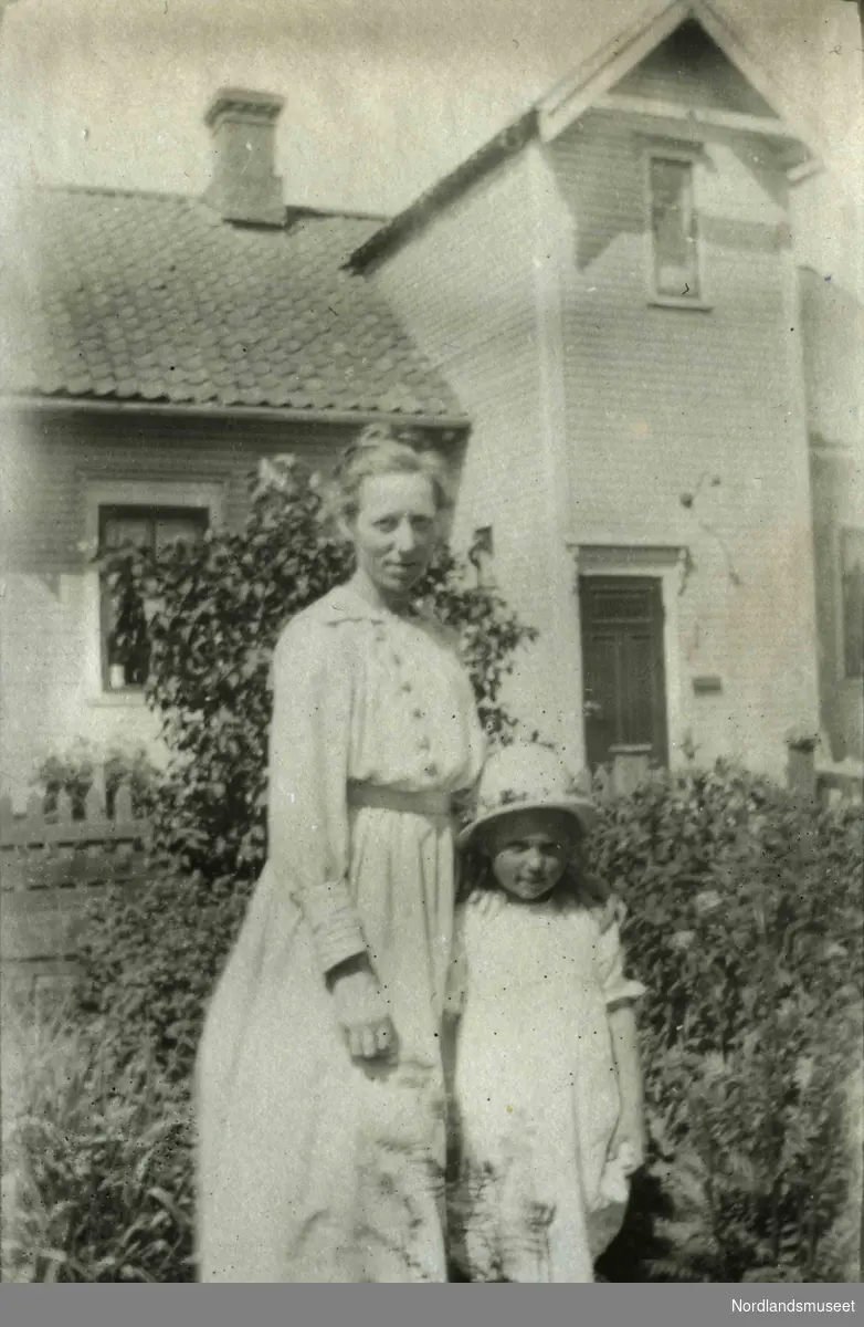 En kvinne og et barn står fremfor et hus. Begge er kledt i lyse klær.