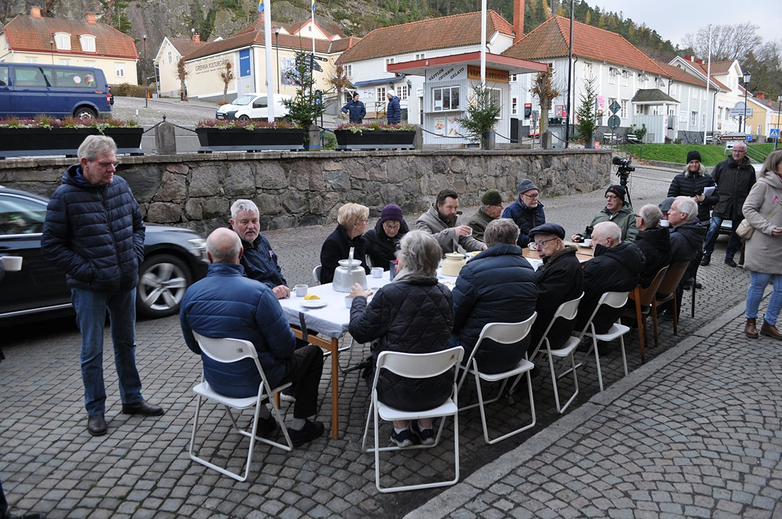 Folk står och sitter vid dukat bord på Brahegatan utanför kafé Fiket i Gränna. Vita plaststolar och en vit pappersduk på fällbordet; en skål med polkagrisar och en julstjärna?