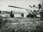 Flygplan Hansa typ 43 / S 3 nr 44 med flottörer står vid vatten, 1925. Sju personer i rörelse vid planet. Mannen i mitten, med vitt hår är Arvid Flory.