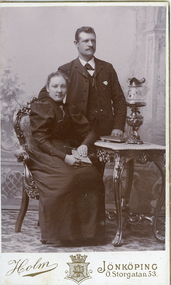 Porträtt av en okänd kvinna och man med mustasch. Mannen står snett bakom kvinnan som sitter vid ett bord. Möjligen ett par. En pokal? på bordet.