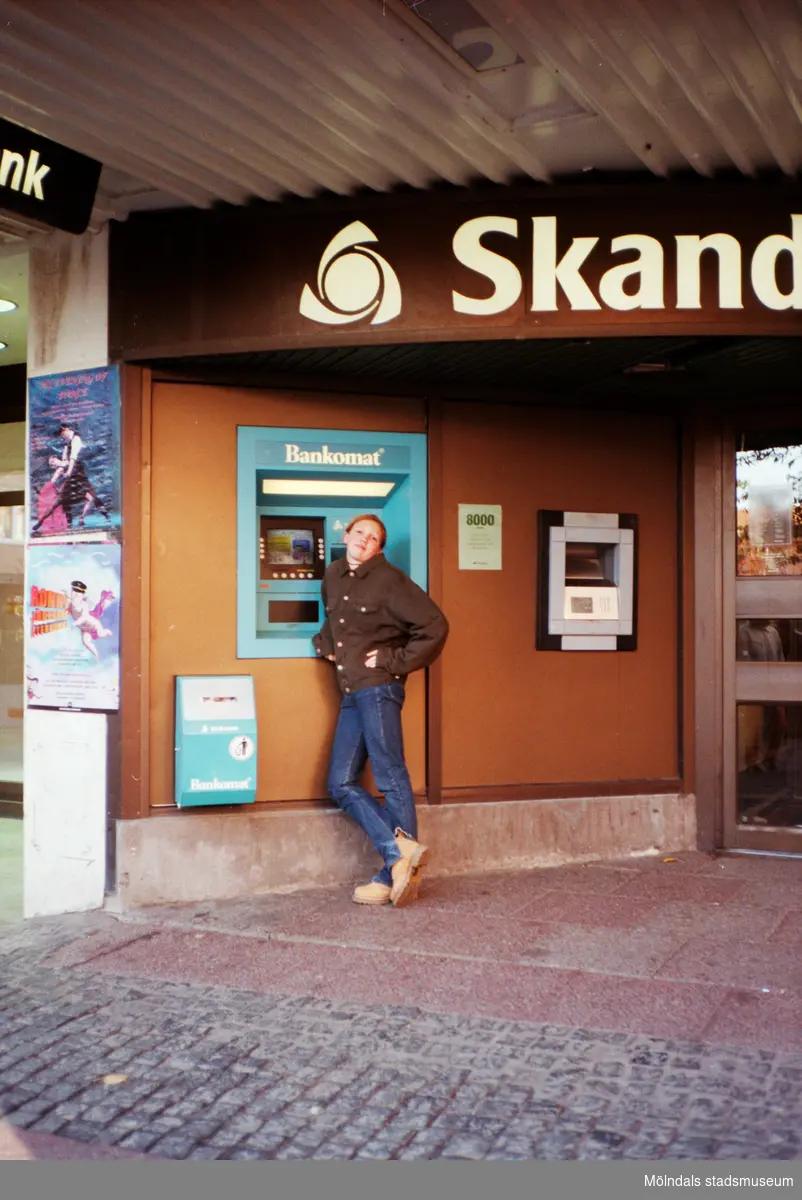 En kille står framför en Bankomat som är placerad utanför Skandinaviska Enskilda banken. Mölndalsbro i dag - ett skolpedagogiskt dokumentationsprojekt på Mölndals museum under oktober 1996. 1996_1061-1076 är gjorda av högstadieelever från Kvarnbyskolan 9C, grupp 3. Se även 1996_0913-0940, gruppbilder på klasserna 1996_1382-1405 samt bilder från den färdiga utställningen 1996_1358-1381.